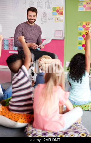 Elementare Schüler heben die Hände zu beantworten Frage als männliche Lehrer liest Geschichte im Klassenzimmer Stockfoto