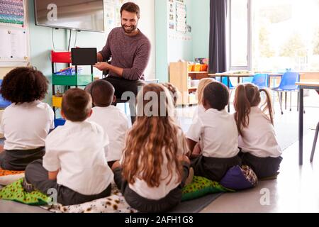 Männliche Lehrer halten Digitale Tablet lehrt Gruppe von Uniformierten Grundschule Schülerinnen und Schüler In der Schule Klassenzimmer Stockfoto