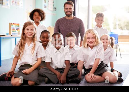 Porträt der Grundschule Schülerinnen und Schüler tragen Uniform sitzen auf dem Boden im Klassenzimmer mit männlichen Lehrer Stockfoto