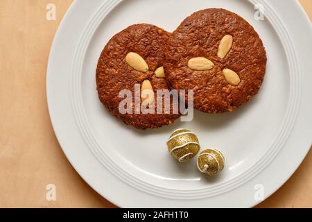 Stillleben mit ein Paar leckeren braunen Elisengingerbreads mit Mandeln und weihnachtliche Dekoration liegen auf einem reinen weißen Porzellanteller auf Holz Stockfoto