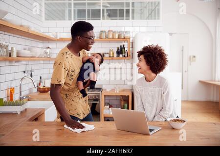 Multi-Tasking Vater hält Baby Sohn und Reinigt die Oberfläche als Mutter verwendet, Laptop und isst Frühstück Stockfoto