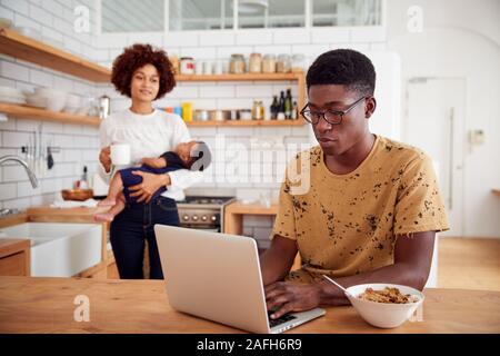 Multi-Tasking Mutter hält Baby Sohn und Macht heißen Getränk als Vater nutzt Laptop und isst Frühstück Stockfoto