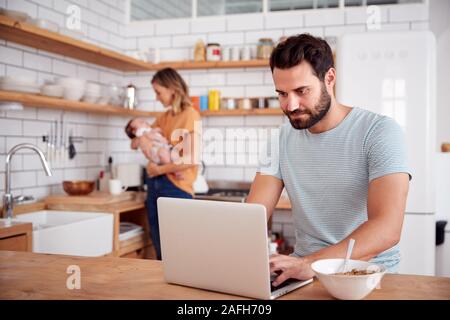 Multi-Tasking Mutter hält Baby Sohn und Macht heißen Getränk als Vater nutzt Laptop und isst Frühstück Stockfoto