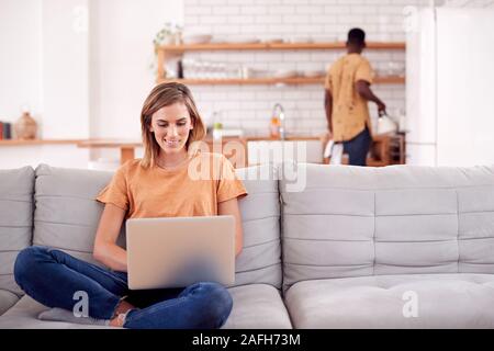 Frau entspannende Sitzen auf einem Sofa zu Hause mit Laptop mit Mann in der Küche hinter Stockfoto