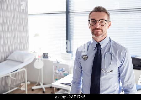Portrait von lächelnden Reife männlicher Arzt mit Stethoskop stehen Schreibtisch im Büro Stockfoto