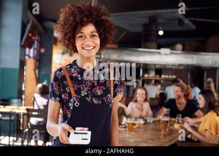 Portrait von Kellnerin Holding Kreditkarte Payment Terminal In belebten Bar Restaurant Stockfoto