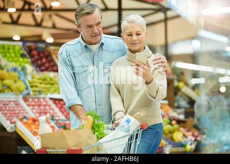 Taille bis Porträt des modernen senior Paar drücken Warenkorb und Überprüfung Liste via Smartphone und genießen Einkaufen im Supermarkt, kopieren Raum Stockfoto