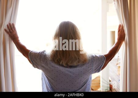 Lächelnd senior weiß Frau den Vorhang zu öffnen, um an einem sonnigen Morgen, Rückansicht, Taille bis Stockfoto