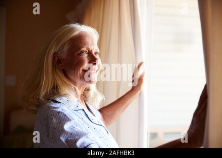 Lächelnd senior weiß Frau den Vorhang zu öffnen, um an einem sonnigen Morgen, Seitenansicht, in der Nähe Stockfoto
