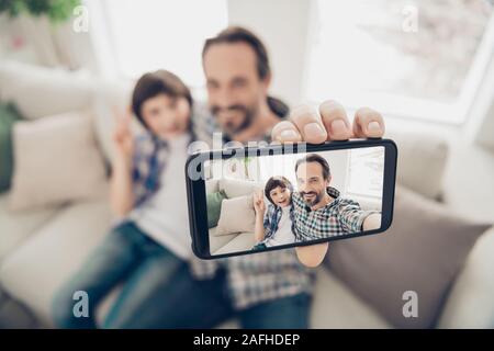 Zwei Ellipsen des Analemmas Portrait von Stattlichen positiv fröhlichen Vater umarmt seinen Sohn umarmen auf der Couch unter selfie auf Telefon angezeigt v-sign in beste Wochenende Stockfoto