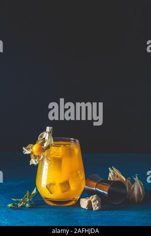Gelb Orange Cocktail mit Mandarine und Rosmarin in Glas mit Wassertropfen Physalis rubro und braunen Zucker auf dunklem Hintergrund mit Bok eingerichtet Stockfoto