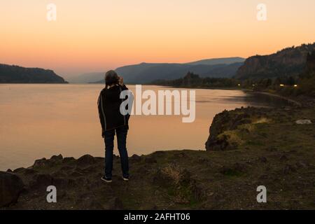 Junge Frau mit Poncho fotografiert den Sonnenuntergang am Corbett Lookout auf dem Columbia River, Oregon Stockfoto