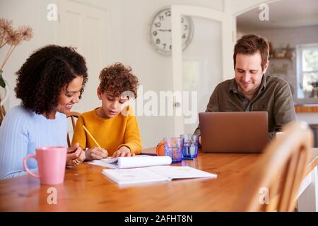 Vater arbeitet auf Laptop als Mutter hilft Sohn mit Heimarbeit auf Tisch Stockfoto