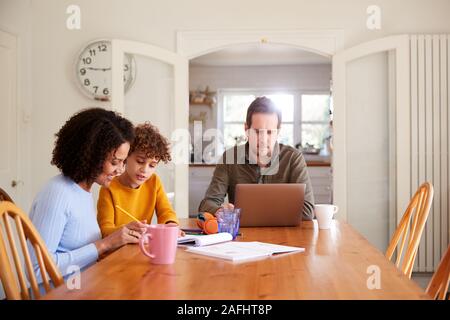 Vater arbeitet auf Laptop als Mutter hilft Sohn mit Heimarbeit auf Tisch Stockfoto
