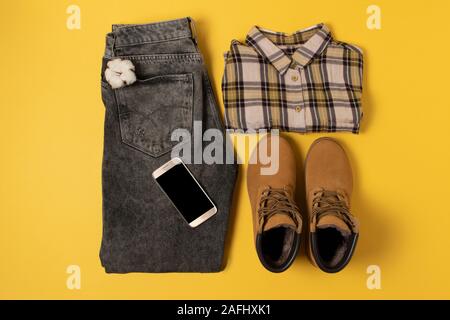 Layout der Herbst Kleidung und Accessoires in warmen Farben auf einem gelben Hintergrund. Jeans, Hemd, Stiefel und Telefon Flat lag in der Nähe Stockfoto