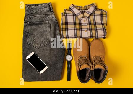 Layout der Herbst Kleidung und Accessoires in warmen Farben auf einem gelben Hintergrund. Jeans, Hemd, Schuhe, Telefon und schauen Sie sich flach in der Nähe Stockfoto