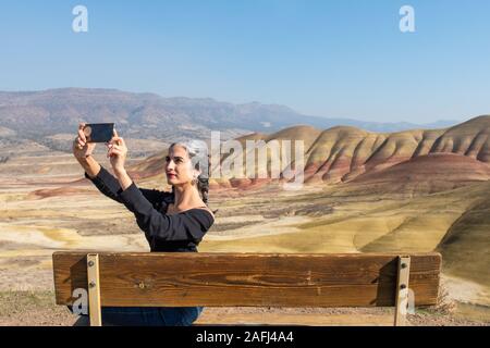 Eine junge Brünette mit grauen Strähnen macht selfies sitzen auf einer Bank in Painted Hills mit Blick auf Stockfoto