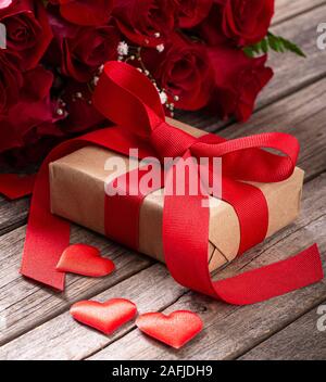 Nahaufnahme von Valentine Geschenk verpackt in braunem Papier und Red Ribbon mit Blumenstrauß aus Rosen auf Holzmöbeln im Landhausstil Oberfläche Stockfoto