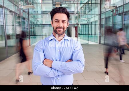 Portrait von Geschäftsmann mit gekreuzten Armen stehen in der Lobby der hektischen modernen Büro Stockfoto