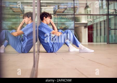 Müde Junge männliche Arzt tragen Scheuert sich gegen Wand im modernen Krankenhaus Gebäude Stockfoto