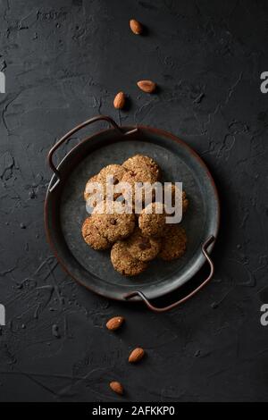 Vegane Spezialitäten Konzept. Almond cookies mit Schokolade Tropfen in Vintage metall Fach und ganze Mandel Muttern auf schwarzem Hintergrund oben Ansichtskopie Raum Stockfoto