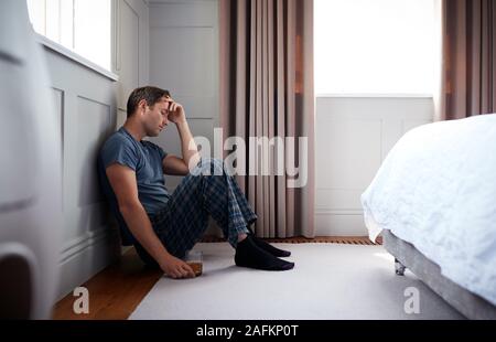 Deprimiert Mann mit Schlafanzug sitzen auf dem Boden des Schlafzimmers Holding Glas Whisky Stockfoto