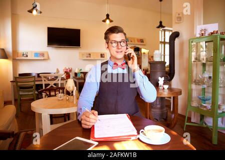 Schuss der Jungen cafe Besitzer Mann mit Bow Tie und Schürze beim Sitzen am Tisch einen Anruf tätigen. Stockfoto