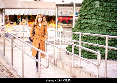 Volle Länge geschossen von Frau mittleren Alters mit Mantel und Tasche beim Gehen aus dem Einkaufszentrum im Winter. Stockfoto