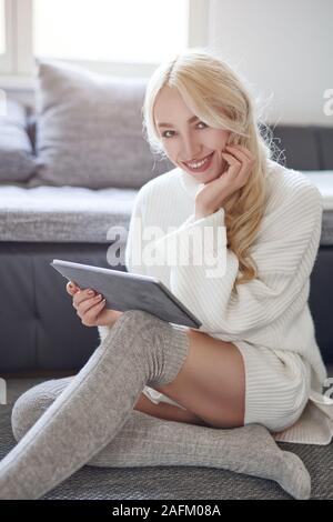 Trendy lächelnde blonde Frau im Winter Leggins und einem warmen Wollpullover zu Hause entspannen mit einem Tablet PC in die Kamera schaut mit einem schönen vivacio Stockfoto