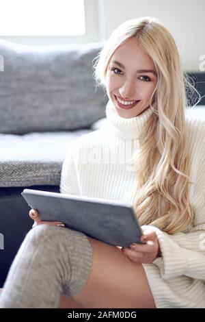 Trendy lächelnde blonde Frau im Winter Leggins und einem warmen Wollpullover zu Hause entspannen mit einem Tablet PC in die Kamera schaut mit einem schönen vivacio Stockfoto