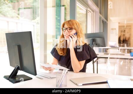 Der Umsatz im mittleren Alter Geschäftsfrau in einem Gespräch ist und dabei einige Schreibarbeit während der Arbeit am Computer im Büro. Stockfoto