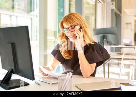 Der Umsatz im mittleren Alter Geschäftsfrau in einem Gespräch ist und dabei einige Schreibarbeit während der Arbeit am Computer im Büro. Stockfoto