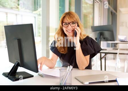 Der Umsatz im mittleren Alter Geschäftsfrau in einem Gespräch ist und dabei einige Schreibarbeit während der Arbeit am Computer im Büro. Stockfoto