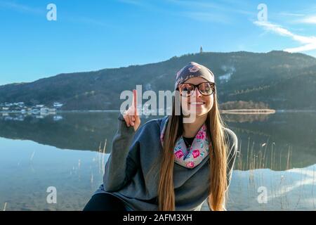 Mädchen mit einem beanie wirft mit Blick auf den See hinter ihr. Ruhige Oberfläche des Sees spiegelt die Berge und den Himmel. Mädchen lächelt, Sie genießen. Stockfoto