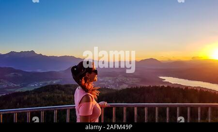 Ein Mädchen steht auf einer Aussichtsplattform und genießen die atemberaubende Aussicht auf das Tal und den Wörthersee während des Sonnenuntergangs. Hügel sind fangen die Stockfoto