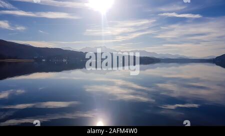 Ein Blick auf den See und die Alpen im Hintergrund. Die ruhige Oberfläche des Sees reflektiert die Berge, Sonne und Wolken. Klare und sonnigen Tag. Ruhe und Re Stockfoto