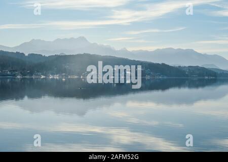 Ein Blick auf den See und die Alpen im Hintergrund. Die ruhige Oberfläche des Sees reflektiert die Berge, Sonne und Wolken. Kleine Stadt am See Stockfoto