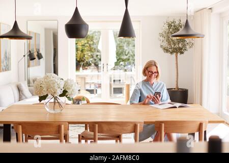 Ältere Frau Entspannende mit Magazin zu Hause auf Handy Sitzen am Esstisch Stockfoto