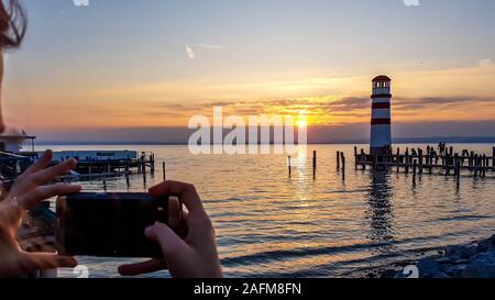 Eine junge Frau, die Bild von einem Leuchtturm auf dem See. Leuchtturm hat weiße und rote Streifen. Sun spiegelt sich im ruhigen Wasser des l Stockfoto