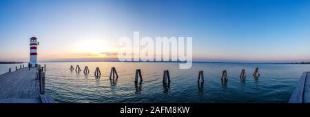 Weiß und Rot gestreifte Leuchtturm im Neusiedlersee, auf österreichisch-ungarischen Grenze. Sonne über dem Horizont, der in den Wolken. Der Himmel ist orange, See Oberfläche Stockfoto