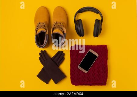 Layout der Herbst Kleidung und Accessoires in warmen Farben auf einem gelben Hintergrund. Handschuhe, Kopfhörer, Stiefel, Telefon und Schal flach in der Nähe. Stockfoto