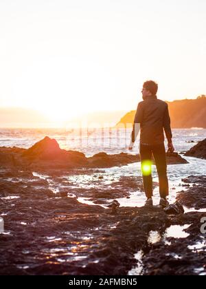 Mann, der auf felsigen Ufer mit Blick auf Meer und Sonnenuntergang Stockfoto