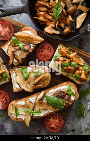 Kochen wilden Wald Pilze. Sandwiches mit gebratenen Pfifferlingen und Steinpilzen, Tomaten und Basilikum auf dunklem Hintergrund der Ansicht von oben Stockfoto