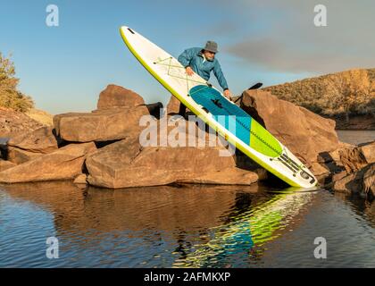 Ältere männliche Paddler stößt eine aufblasbare Stand up paddleboard von einem felsigen Ufer des Bergsee - horsetooth Reservoir in Northern Colorado Stockfoto
