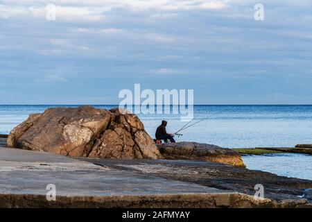 Ein einsamer Fischer mit Angel sitzt auf einem Felsen am Meer. Aktiver Urlaub am Schwarzen Meer. Stockfoto