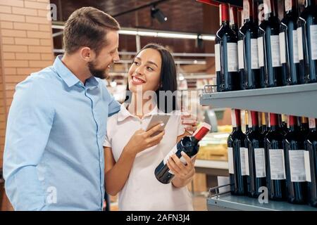 Die täglichen Einkäufe. Paar im Supermarkt zusammen an Alkohol Abteilung umarmen Auswahl Wein Frau surfen Smartphone betrachten einander glücklich Stockfoto