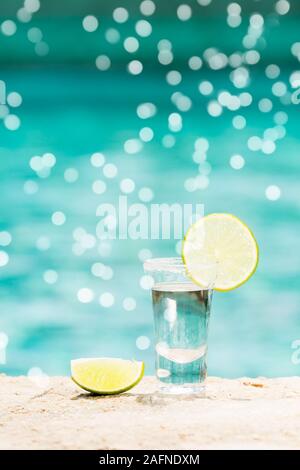 Ein Tequila, Schüsse mit in Scheiben geschnittenen Kalk an den Strand. Transparente Alkohol. Heraus hängen. Vertikale mit sanften Festliche bokeh Stockfoto