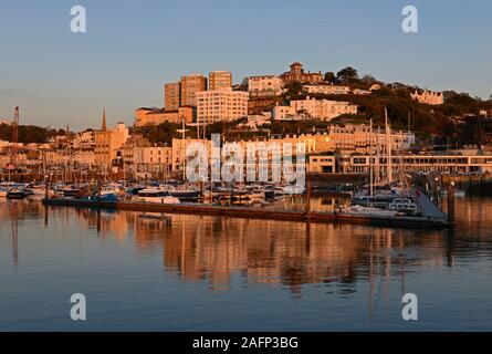 Jachten und andere Yachten in der Marina bei Torquay, Devon, UK festgemacht, wie die Sonne, die Stadt hinter die Beleuchtung mit Orange. Stockfoto
