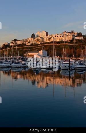 Jachten und andere Yachten in der Marina bei Torquay, Devon, UK festgemacht, wie die Sonne, die Stadt hinter die Beleuchtung mit Orange. Stockfoto