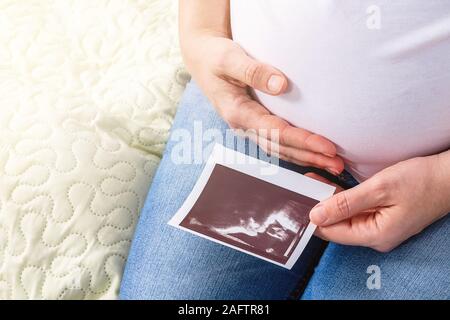 Schwangere Frau saß auf dem Bett. Eine Hand schmiegt sich an ihren Bauch und in anderen hält Ultraschall Foto der Zukunft Baby. Schwangerschaft Konzept. Dritten Trimester. Babys f Stockfoto